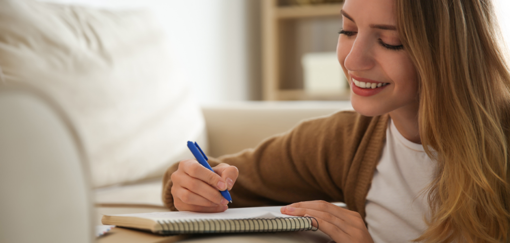 A happy woman writing a letter on a sofa at home.
