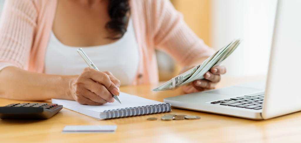 A woman managing her family's budget, writing in a notebook, holding dollar bills, and using a laptop and calculator.