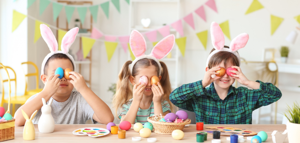 Three kids with bunny ears are sitting at a table, happily painting Easter eggs.