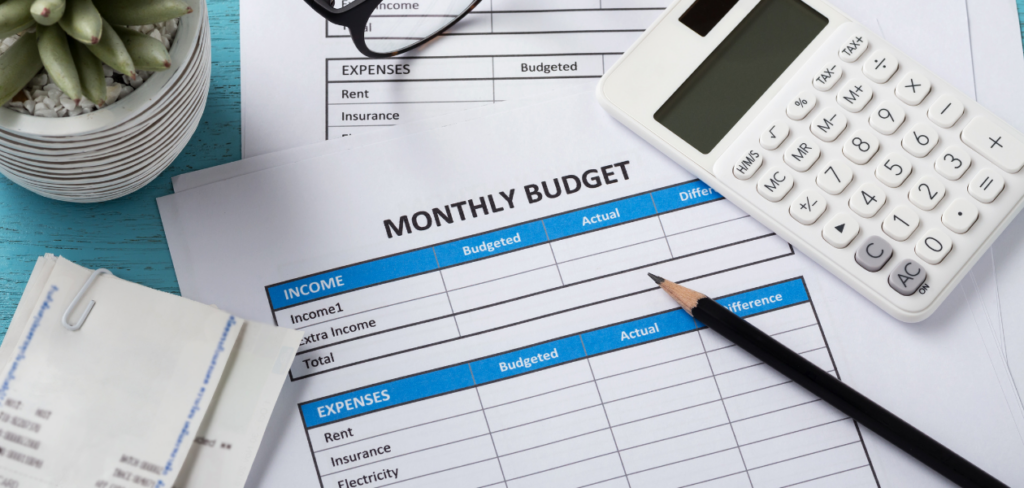 Monthly budget planning with a white calculator, pencil, eyeglasses, receipts, and a monthly budget sheet on a blue desk.
