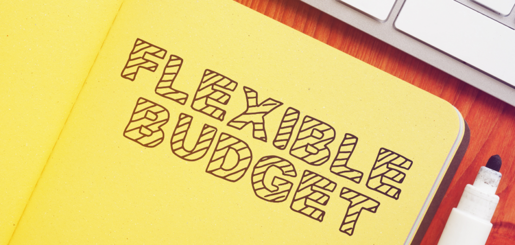"Flexible budget" is written on a yellow notebook with a marker, alongside a keyboard, on a wooden table.
