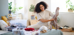 A smiling woman sitting on her bed while sorting and organizing clothes into donation bins, surrounded by labeled containers.