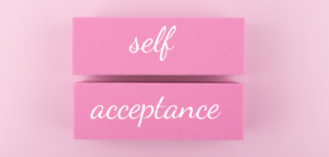 Two pink blocks stacked on a pastel pink background, with the words 'self' and 'acceptance' written in white script.
