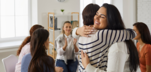 Two women warmly embracing in a supportive group setting, with others around them clapping and smiling.