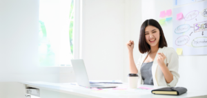 A young woman sitting at a desk, smiling and celebrating success with clenched fists, in a bright office space.