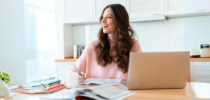 Woman smiling while sitting at a table with a laptop, books, and a cup of coffee.