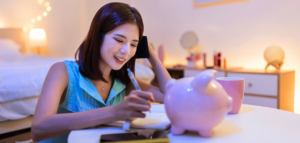 Young woman sitting at a table, smiling while managing finances — writing in a notebook, talking on the phone, with a pink piggy bank and coffee cup nearby.