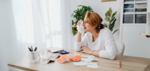 Woman sitting at a desk with bills, receipts, and cash, holding a tissue to her face and appearing stressed while budgeting or managing finances.