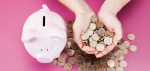 A pink piggy bank next to a large pile of coins with hands holding a handful of them, set against a bright pink background.
