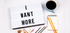 A lightbox sign that reads 'I WANT MORE,' a cup of coffee, notebook, pencils, colorful paperclips, and wooden stamp pieces.