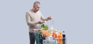 Man looking shocked at a long grocery receipt while standing next to a full shopping cart.