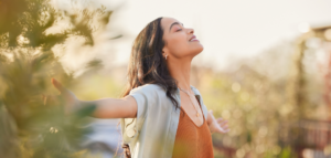 A woman standing outdoors with her arms open wide, eyes closed, and a peaceful smile.