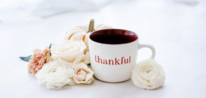 A white mug with "thankful" written on it, surrounded by flowers and a small pumpkin.