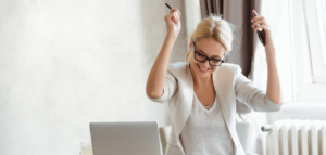A joyful woman sitting at her desk with a laptop, smiling and raising her arms in celebration.