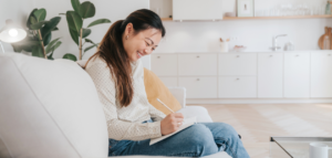 A smiling woman sitting on a couch, writing in a notebook