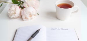 A table featuring a gratitude journal, a pen, a cup of tea, and soft pink peonies.