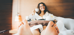 A happy woman enjoying breakfast in bed, wearing a white bathrobe and smiling while holding a cup of coffee or tea, with a tray of breakfast items in front.