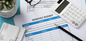 A flat lay of a 'Monthly Budget' worksheet surrounded by a calculator, pencil, eyeglasses, receipts, and a small potted plant.