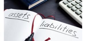Close-up view of an open notebook with the words "assets" and "liabilities" written on facing pages, a black marker and a calculator.