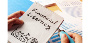 Close-up of hands holding a spiral notebook with the words "Financial Literacy" written on it. A blue marker is also visible, along with some financial charts and graphs in the background.