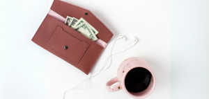 A mauve leather wallet with cash, white earbuds, and a pink coffee mug on a white background.