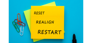 A yellow sticky note on a blue background with the words "RESET," "REALIGN," and "RESTART" written on it. A black marker and paper clips are also visible.