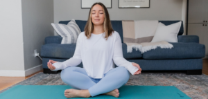 A woman meditates on a yoga mat in front of a couch.