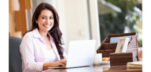 A smiling woman sits at a desk, working on a laptop. A mug and family photos are visible on the desk.