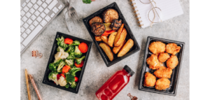 A flat lay of a lunchbox setup featuring three plastic containers filled with food, a bottle of juice, and everyday desk items like a keyboard, pens, and glasses.