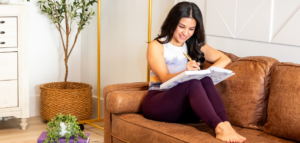 A woman sits on a brown leather couch, smiling as she writes in a large notebook.