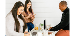 Three women collaborate at a table, working on laptops and notebooks. There are coffee cups and pastries on the table.