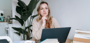 A woman sits at a desk, thoughtfully looking off to the side, with a laptop in front of her and a stack of books beside it.