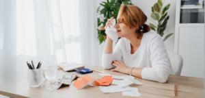 A woman sits at a table, visibly upset, surrounded by bills, cash, and a calculator. She holds a tissue to her face.