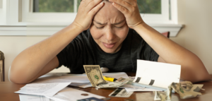 A woman sits at a table, head in hands, visibly distressed, surrounded by bills, cash, and credit cards.