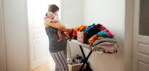 A woman in pajamas holds a sleeping baby on her shoulder while sorting through a large pile of laundry on an ironing board.