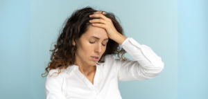 Close-up of a woman holding her head in her hand, eyes closed, appearing stressed or overwhelmed against a light blue background.