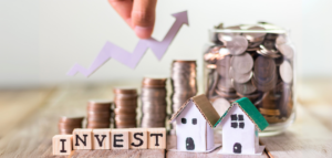 Wooden blocks spelling "INVEST" are in front of stacks of coins, miniature houses, and a jar of coins. An upward-pointing arrow is above the coins.