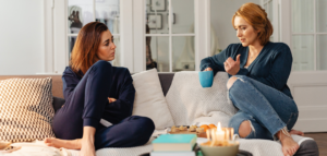 Two women sit on a couch, one appearing concerned while the other speaks, holding a mug. A candle and small plate of food are on the coffee table between them.