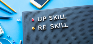 A letterboard on a blue background displays the words "UP SKILL" and "RE SKILL" in white letters. Office supplies like pens, pencils, a smartphone, and pushpins surround the letterboard.