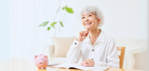 A smiling older woman sits at a table, looking thoughtfully off to the side. A pink piggy bank and a cell phone are on the table in front of her.