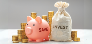 A pink piggy bank with the word 'SAVE' printed on it and a burlap sack with the word 'INVEST' sit in front of several stacks of gold coins.