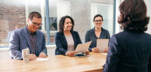 Three people sit at a table during a job interview, smiling and looking at the interviewee.