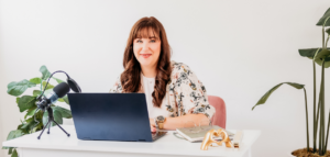 A woman smiles at the camera while sitting at a desk with a laptop and notebooks.