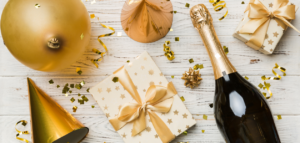 A bottle of champagne, gift boxes, balloons and confetti on a white wooden table.