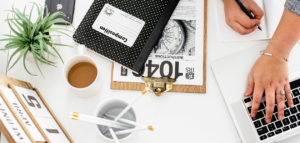 A flatlay image of a workspace showing a laptop, a clipboard with paperwork, a notebook, a small succulent plant, a cup of coffee, a desk calendar, and pencils in a holder.