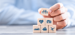 A hand places a wooden block with a family icon on top of a stack of blocks featuring a house, a heart, a car, a piggy bank, and an airplane, representing insurance products.