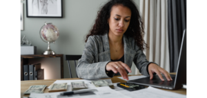 A woman sits at a wooden desk, concentrating on her laptop. Spread out on the desk around her are papers, a calculator, and stacks of cash.