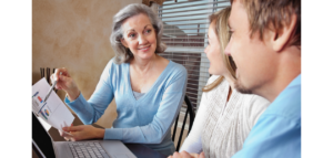 A woman sits at a table with a young couple, pointing to a chart on a piece of paper as she explains something to them. A laptop is on the table in front of them.