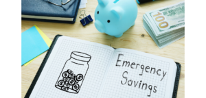 A notebook open to a page with the words "Emergency Savings" written on it, next to a drawing of a jar filled with coins. A piggy bank, cash, and other office supplies are also visible on the desk.