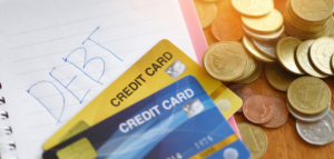 A close-up shot of a notebook with the word "DEBT" handwritten on it, alongside two credit cards and a pile of coins.